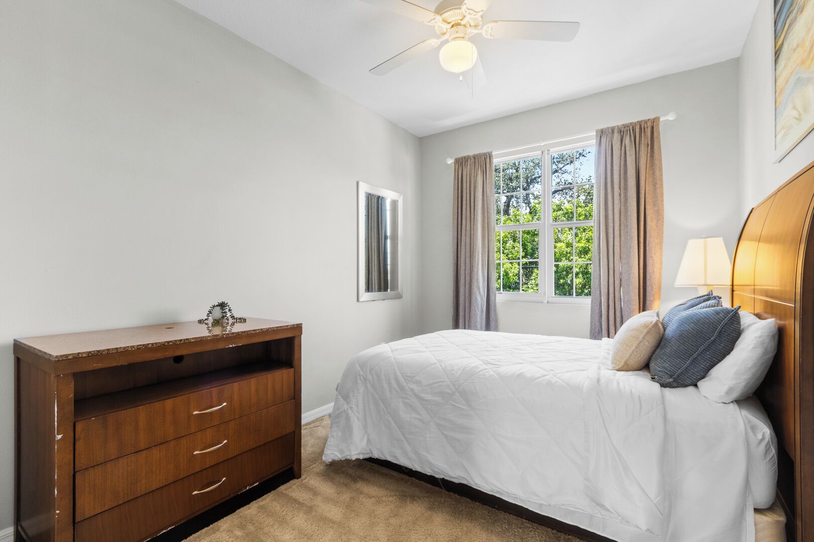 A bedroom with a bed, a wooden dresser, a ceiling fan, and a window with curtains.