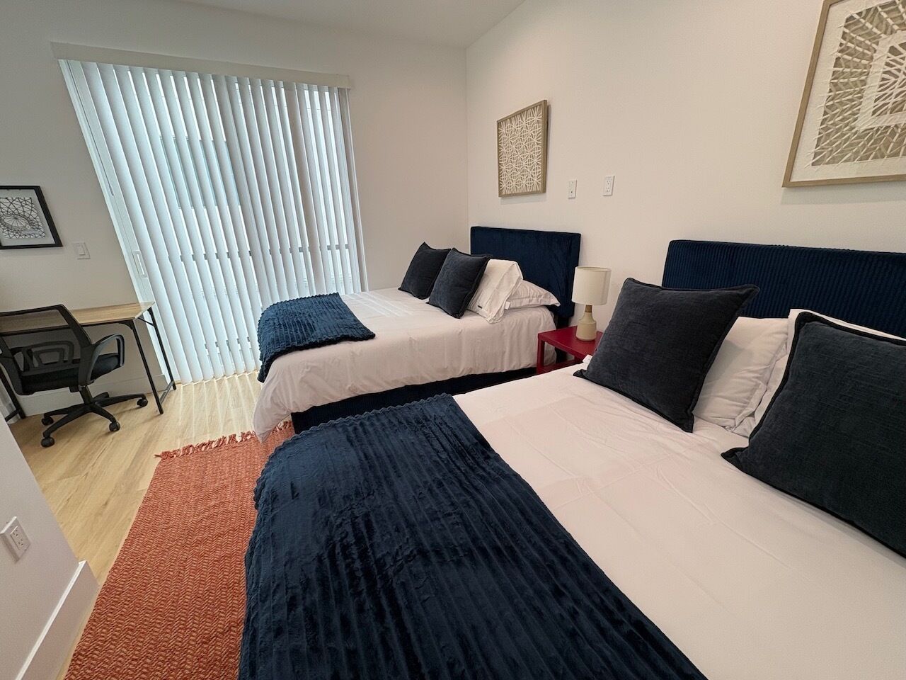 A hotel room with two beds, a desk, and a window with blinds.