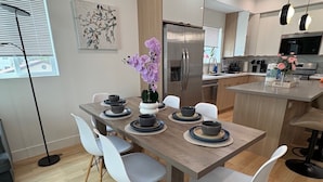Uma cozinha moderna com geladeira de aço inoxidável, uma mesa de jantar com cadeiras brancas e um vaso com flores roxas.