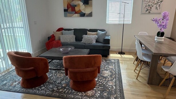 Uma sala de estar moderna com um sofá, duas poltronas, uma mesa de centro e uma mesa de jantar com cadeiras. Há um quadro abstrato grande na parede e um vaso com flores sobre a mesa de jantar.