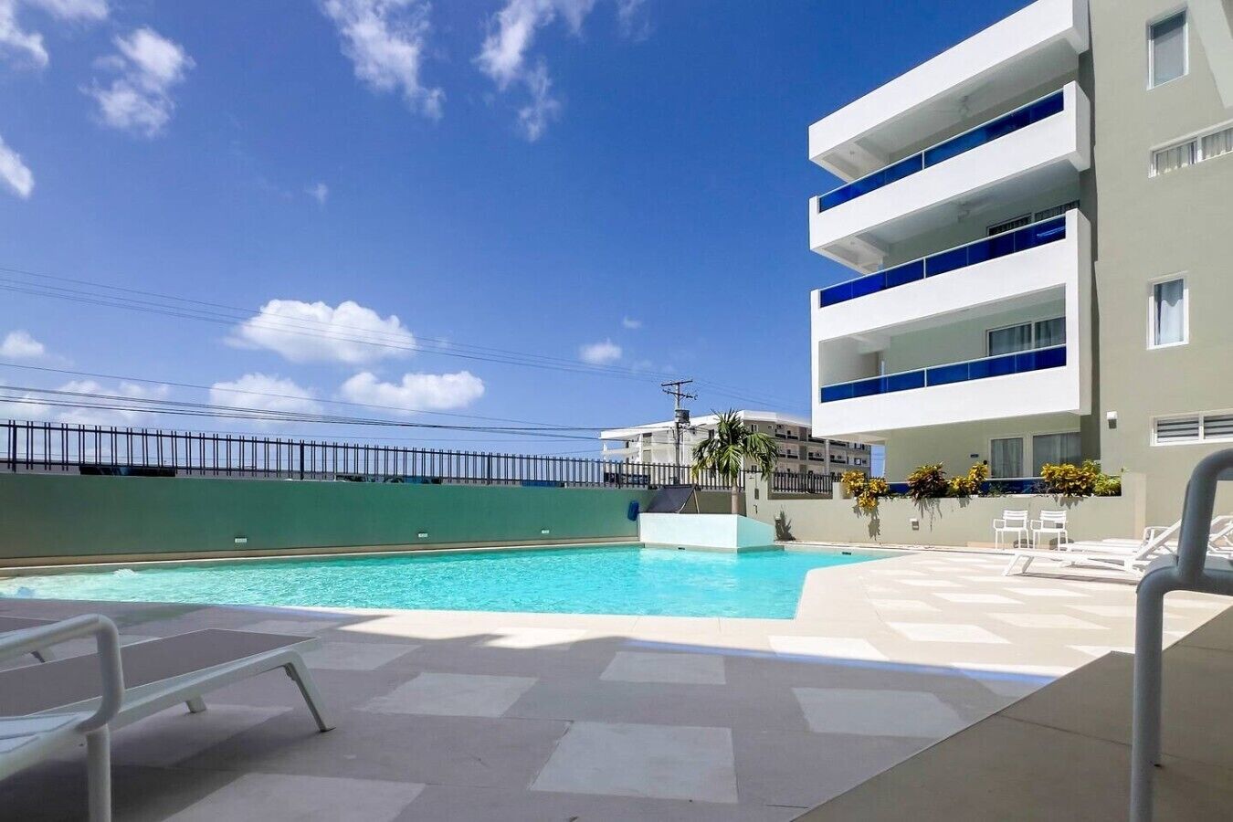 Zona de piscina con cielo despejado, un edificio con balcones y una cerca.