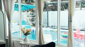 A room with a large window, a black sofa, a small round table with a vase, and a view of a swimming pool with inflatable toys. - Skylux Ocean House: 3BR Private Pool, Game Room & Cinema-Size TV (Hollywood)