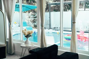 A room with a large window, a black sofa, a small round table with a vase, and a view of a swimming pool with inflatable toys. - Skylux Ocean House: 3BR Private Pool, Game Room & Cinema-Size TV (Hollywood)