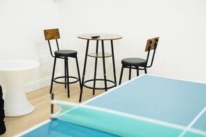 Une pièce comprenant une table de ping-pong, deux chaises, une petite table ronde et une table sur pied, de couleur blanche.
