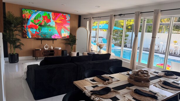 A modern living room with a black sofa, a flat-screen TV, and a dining area with a wooden table and woven placemats. - Skylux Ocean House: 3BR Private Pool, Game Room & Cinema-Size TV (Hollywood)