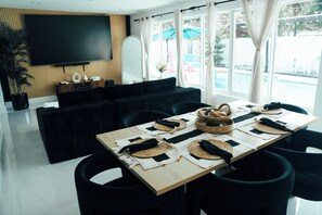 A modern living room with a large flat-screen TV, dark sofas, a wooden accent wall, a dining area with a wooden table set for four, and a view of a pool outside. - Skylux Ocean House: 3BR Private Pool, Game Room & Cinema-Size TV (Hollywood)