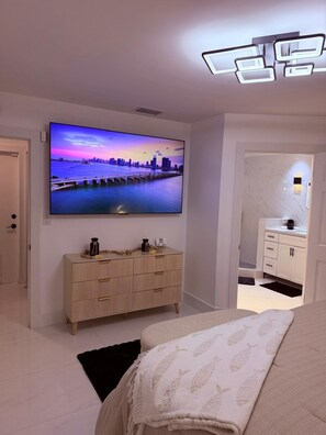 A modern bedroom with a large TV, a wooden dresser, and a white bed. - Skylux Ocean House: 3BR Private Pool, Game Room & Cinema-Size TV (Hollywood)