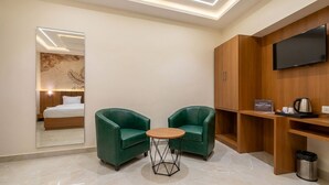 A hotel room with a bed, two green armchairs, a small round table, a TV mounted on the wall, and a wooden cabinet with a kettle on top. - RAS VILAS PALACE - Beside Prem Mandir, Vrindavan (Vrindavan)