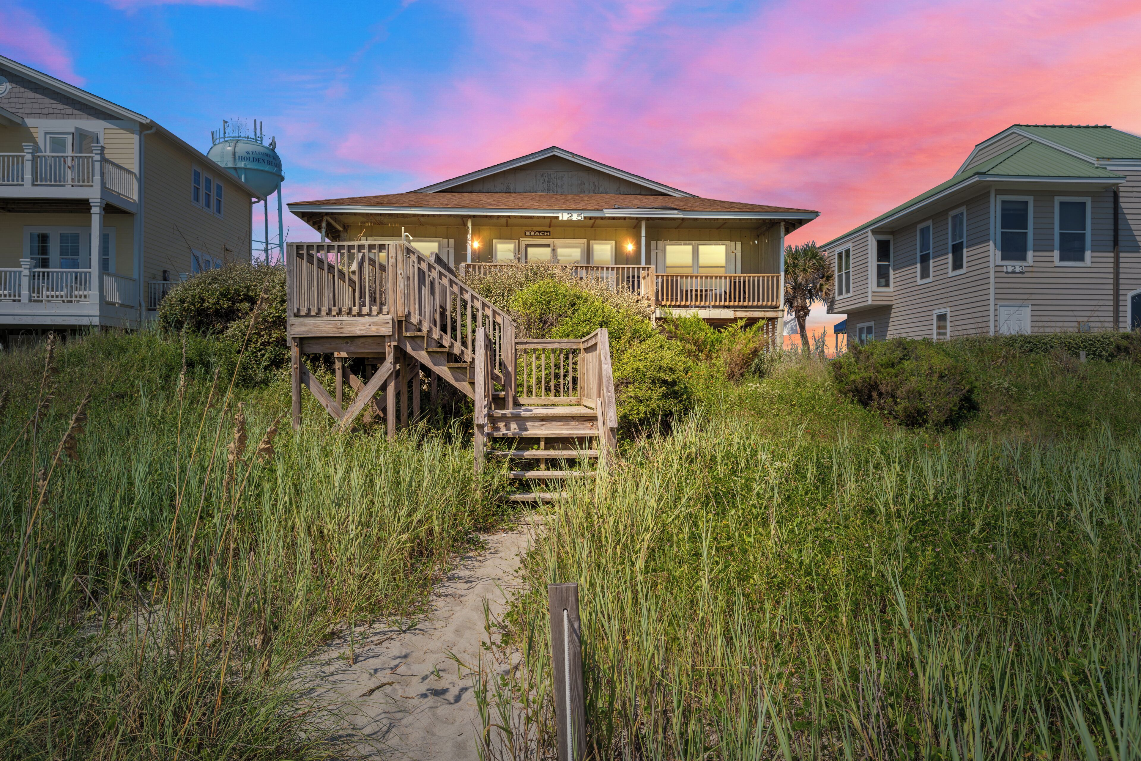 A beachfront hotel with a wooden staircase leading to a balcony, surrounded by tall grass and sand.