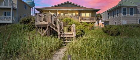 A beachfront hotel with a wooden staircase leading to a balcony, surrounded by tall grass and sand.