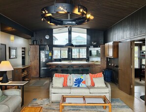 A modern kitchen with a central island, a dining area with a sofa and chairs, and a large window providing a view of the outside. - Oceanview 3BR, 2BA Holden Beach retreat with WiFi and full kitchen (Holden Beach)