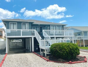 A two-story beach house with a covered entrance, a small garden, and a clear blue sky. - 3BR Holden Beach retreat with private pool (Holden Beach)