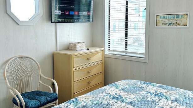 Un dormitorio con cama, una silla, cómoda, televisor y ventana.