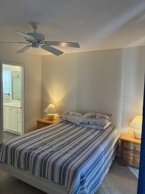 Un dormitorio con cama, ventilador de techo, mesita de noche con lámpara y puerta que da a un baño.