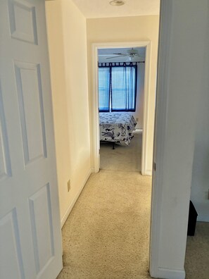 Un pasillo que conduce a un dormitorio con una cama, ventilador de techo y ventana con persianas.