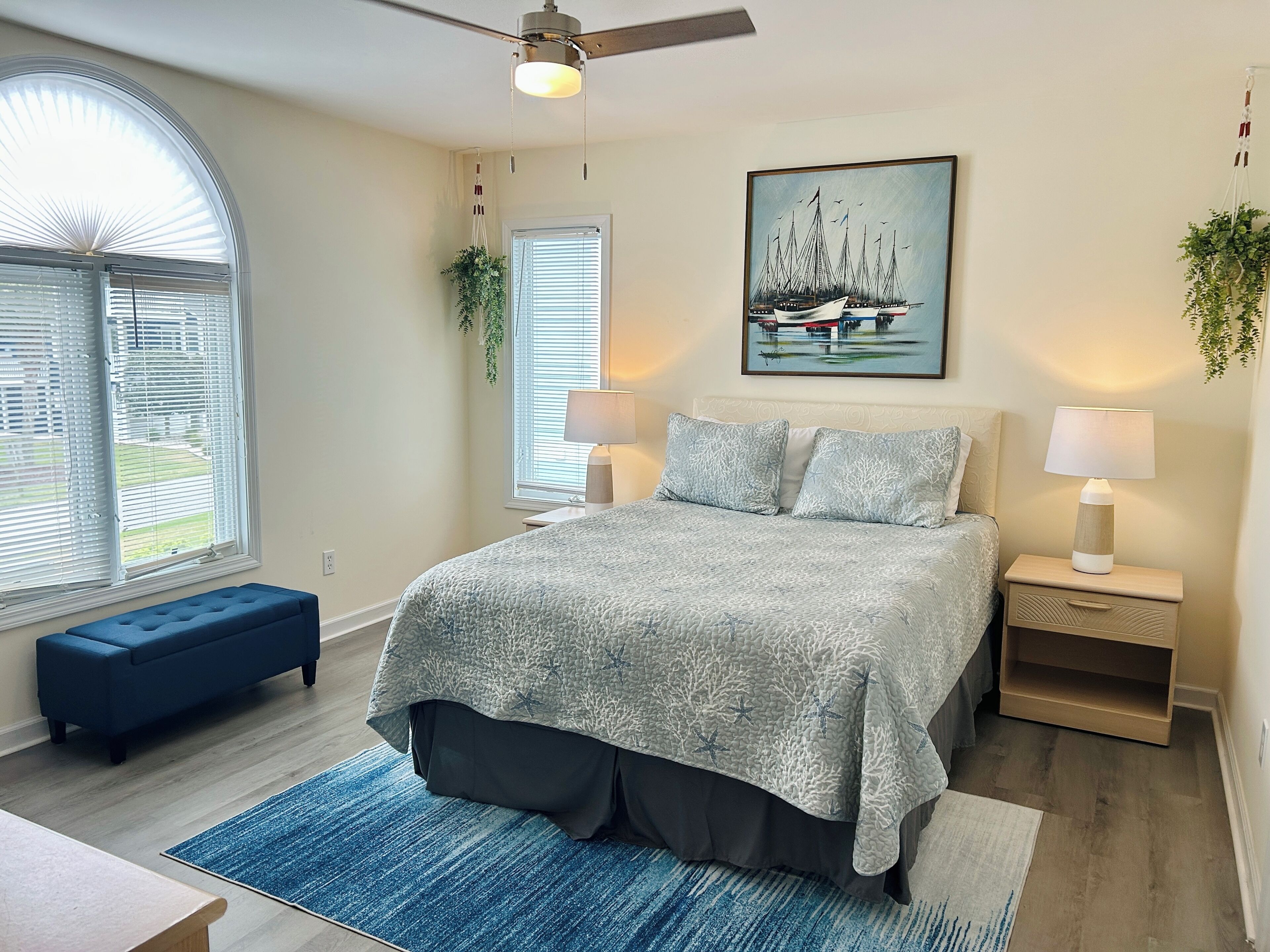 A bedroom with a bed, bedside tables, a ceiling fan, a window with blinds, a blue bench, and a painting on the wall.