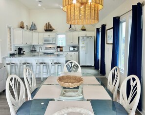 A modern kitchen with white cabinets, a central island, bar stools, a dining table with placemats, and a wicker centerpiece. - 4BR retreat with dock overlooking the ICW (Holden Beach)