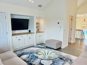 A modern living room with a flat-screen TV, white cabinetry, a glass coffee table, and a view to the dining area. - 4BR retreat with dock overlooking the ICW (Holden Beach)
