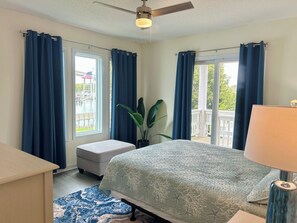 Ein Schlafzimmer mit Bett, Schreibtisch, Stuhl, einem Fenster mit Vorhängen, Deckenventilator und Blick auf einen Balkon.
