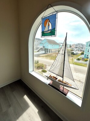 A room with a large arched window overlooking a residential area, a small sailboat model, and a stained glass window decoration. - 4BR retreat with dock overlooking the ICW (Holden Beach)