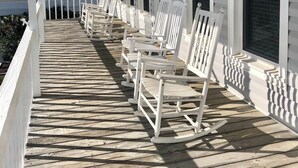 A covered porch with white rocking chairs aligned along the wall, a ceiling light fixture, and windows with blinds. - 4BR, 2BA Holden Beach retreat located on the 2nd row (Holden Beach)