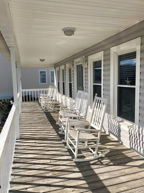 A covered porch with white rocking chairs aligned along the wall, a ceiling light fixture, and windows with blinds. - 4BR, 2BA Holden Beach retreat located on the 2nd row (Holden Beach)