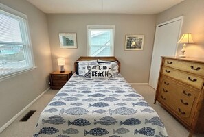 Ein Schlafzimmer mit Bett, Nachttisch, Kommode und zwei Fenstern mit Jalousien.
