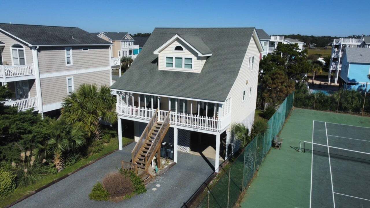 A two-story house with a balcony, a staircase leading to a deck, and a tennis court in the background.