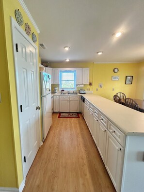 A kitchen with white cabinets, a window with blinds, a sink, and a dining area with chairs. - "Courtside" cozy 4 bedroom home, short walk to the Beach!! (Holden Beach)