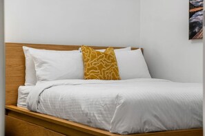 A bed with white bedding and a single patterned pillow, a wooden headboard, and a plain wall with a framed picture. - Sage & Saddle | Balcony | UG Park | Beltline | BBQ (Calgary)