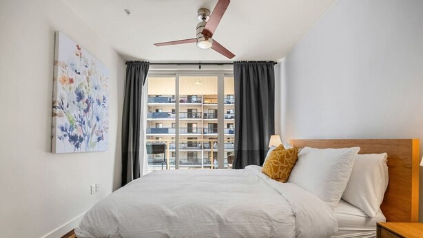 Quarto de hotel com cama, ventilador de teto, janela com cortinas e vista para um edifício.
