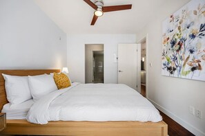 Un dormitorio con una cama de madera, ropa de cama blanca, un ventilador de techo y una obra de arte floral en la pared.