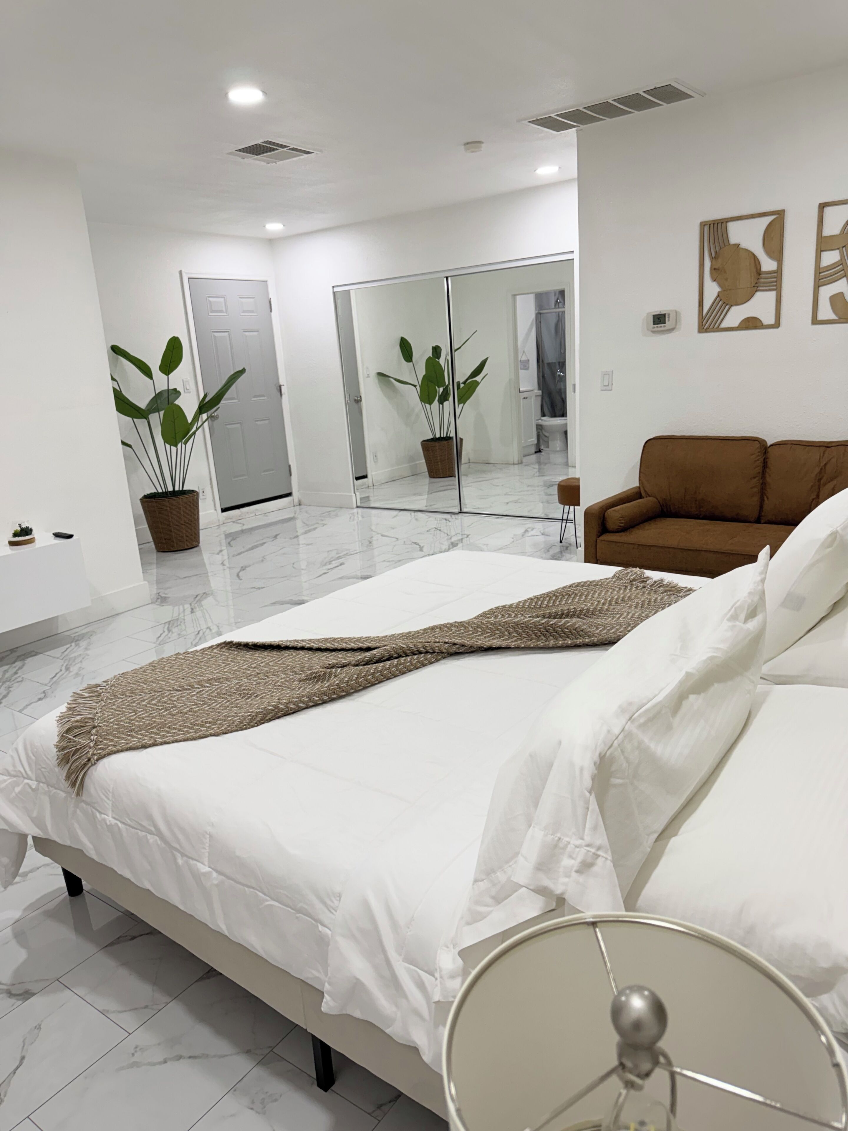 Un dormitorio con una cama, un sofá marrón, una planta en maceta, un espejo y un televisor instalado en la pared.
