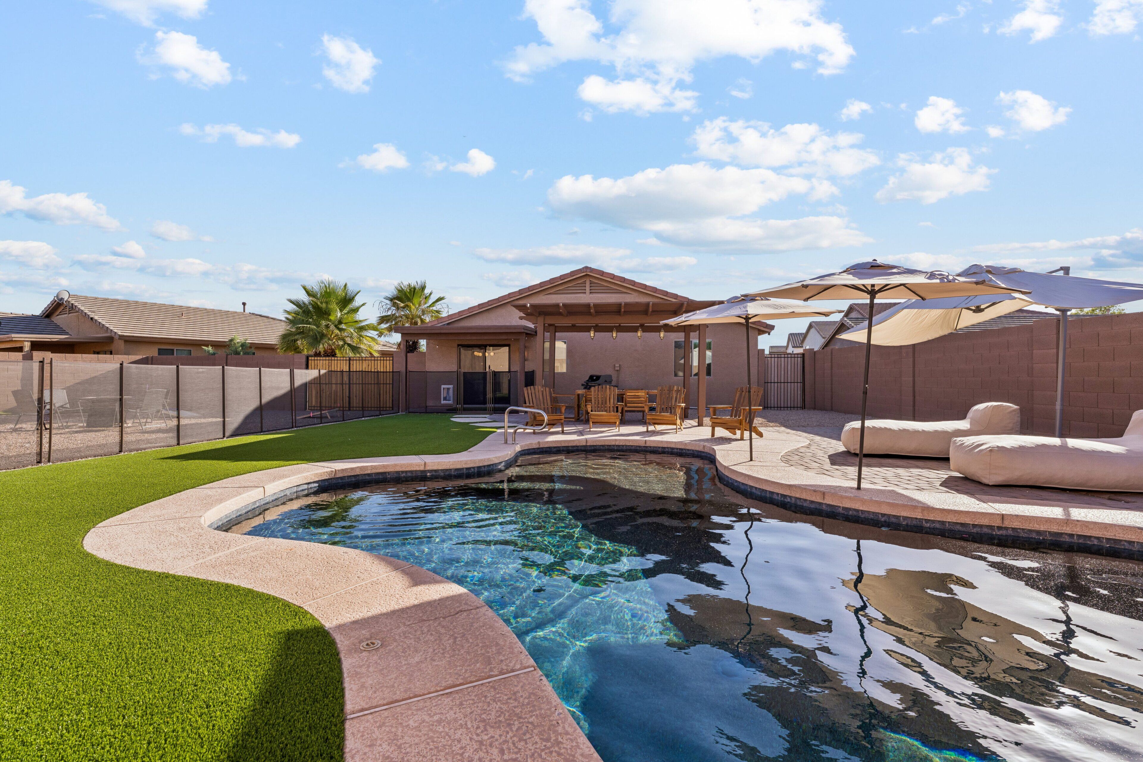 Une piscine à bord incurvé, entourée d’une clôture, et un espace patio aménagé avec des chaises et des parasols.