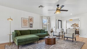 A modern living room with a green sofa, a patterned rug, and a ceiling fan. - Oasis on Aubrac | Modern 5BR | Private Pool | Pergola & Firepit | Sleeps 8 (San Tan Valley)