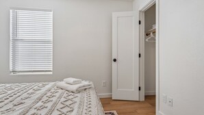 A bedroom with a bed, a window with blinds, a door leading to a closet, and a wooden floor. - Oasis on Aubrac | Modern 5BR | Private Pool | Pergola & Firepit | Sleeps 8 (San Tan Valley)