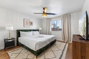 Habitación de hotel con cama, ventilador de techo, televisor, mesita de noche, lámpara y ventana con cortinas.