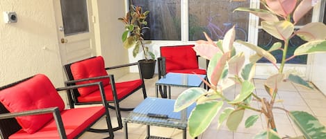 A sunlit patio with red cushioned chairs, a small table, and potted plants.