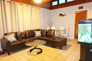 A living room with a brown leather sectional sofa, a glass coffee table, a dining table with chairs, and a fish tank. - Charming 2-bedroom house on the Space Coast. (Melbourne)