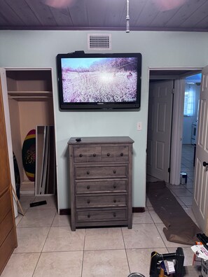 A living room with a television mounted on the wall, a wooden dresser, and a hallway leading to another room. - Charming 2-bedroom house on the Space Coast. (Melbourne)