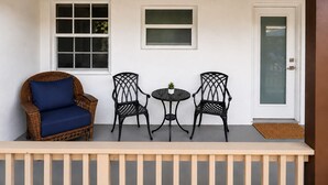 Een veranda met een rieten stoel, twee metalen stoelen, een kleine tafel en een deur.