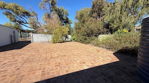 A paved backyard with a fence, trees, and a building in the background.