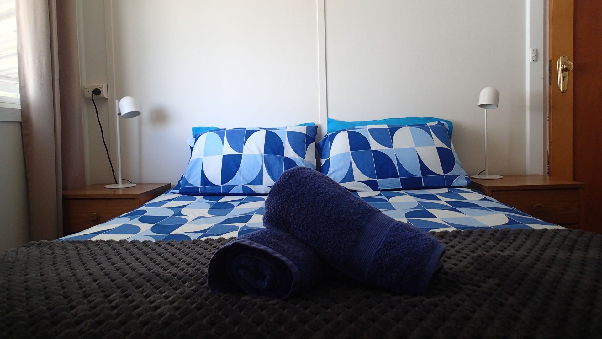 Een opgemaakt bed met een geborduurde sprei, twee kussens en een opgerolde handdoek op een getextureerd vloerkleed.