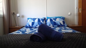 Una cama bien hecha con una colcha estampada, dos almohadas y una toalla enrollada sobre una alfombra con textura.