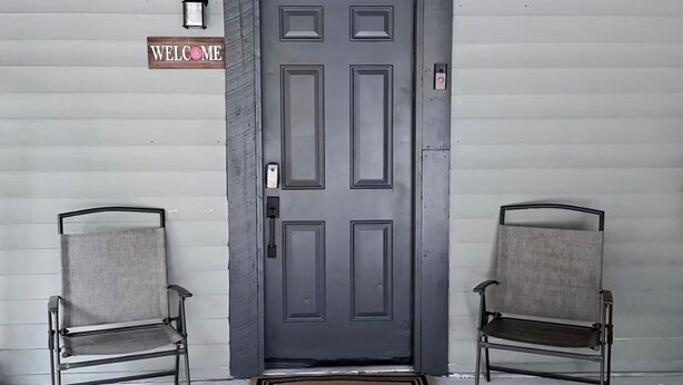 Uma porta cinza com a placa 'WELCOME', duas cadeiras de metal e um tapete de entrada.