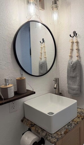 A bathroom with a round mirror, a white sink, a towel rack with two towels, and a soap dispenser. - Cozy Private Studio • 6 Min to Busch Gardens (Tampa)