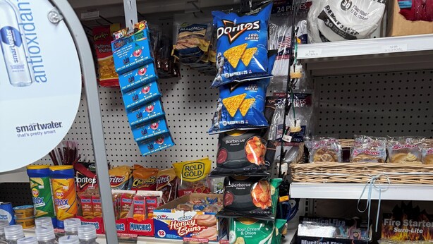 Hylly, joka on täynnä erilaisia välipaloja ja juomia, kuten Doritos-sipsejä, Smartwater-vesiä ja muita pakattuja elintarvikkeita.