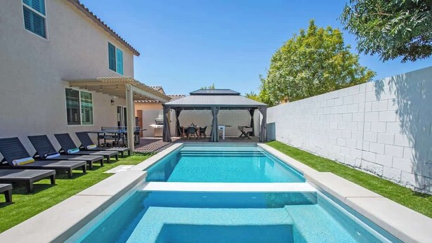 Una piscina rectangular con un área de estar cubierta y sillas reclinables.