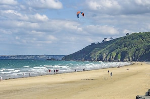 Ein Strand mit Menschen, die am Ufer entlanggehen, einen Drachen am Himmel und eine Hügelkette mit Bäumen im Hintergrund.