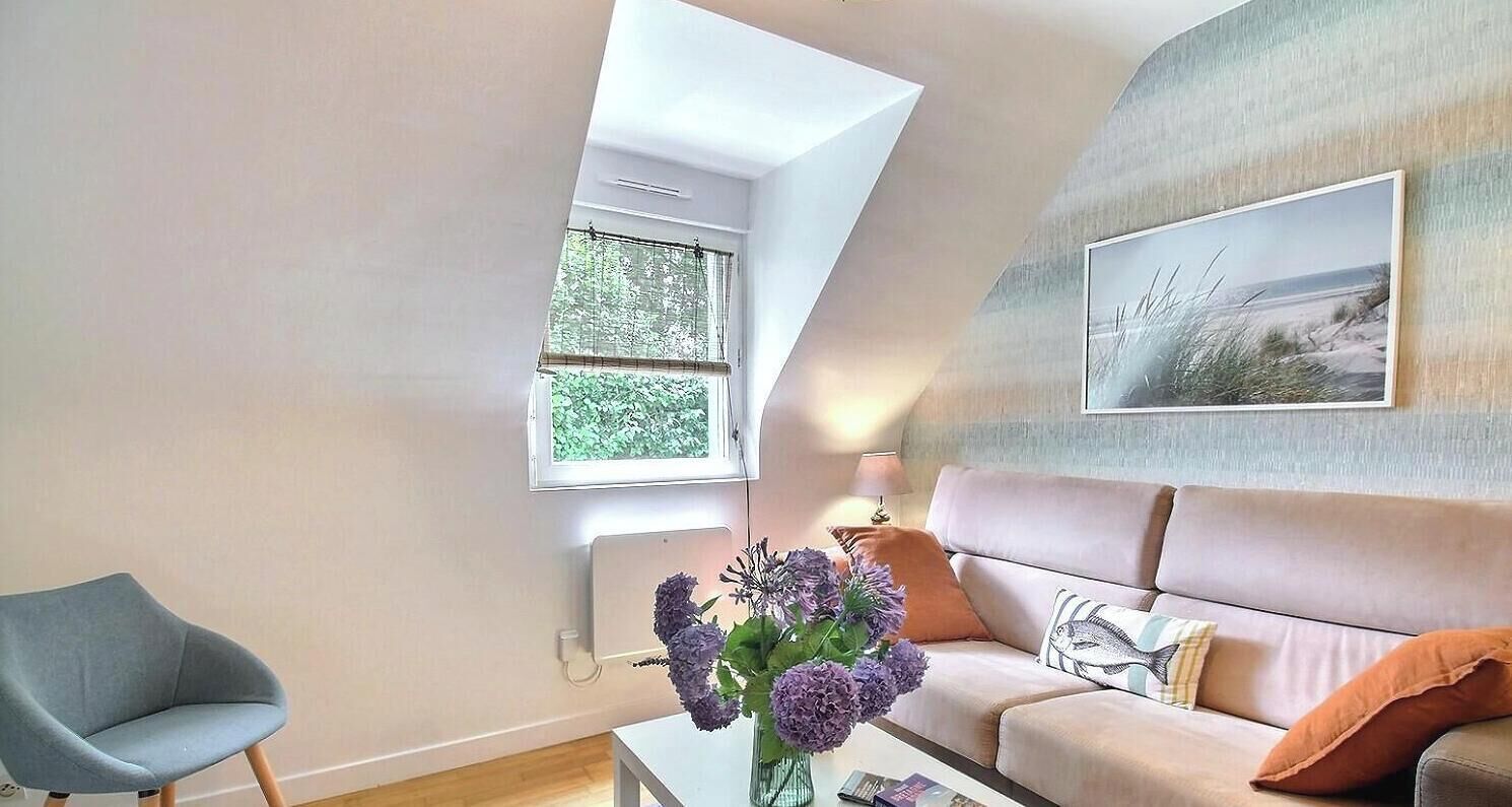 A living room with a sofa, a chair, a table with a vase of flowers, a window, and a wall-mounted picture.